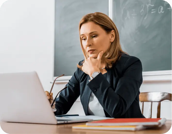 Educator working on a laptop in a classroom, reviewing information while seated at a desk with a chalkboard in the background.