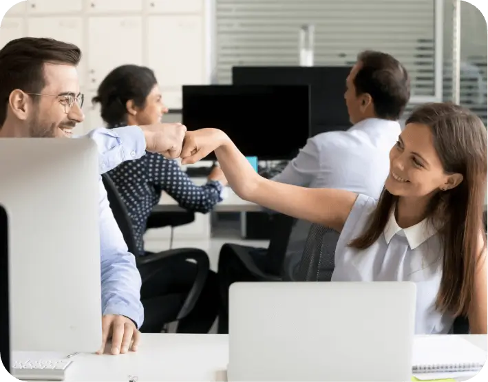 Coworkers celebrating a win with a fist bump while working at laptops in a modern office environment.